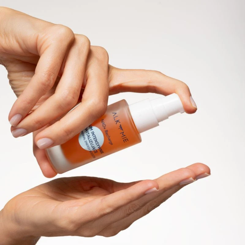 Dłonie z pomarańczowym serum NAD+ Recharge ALKMIE na jasnym tle